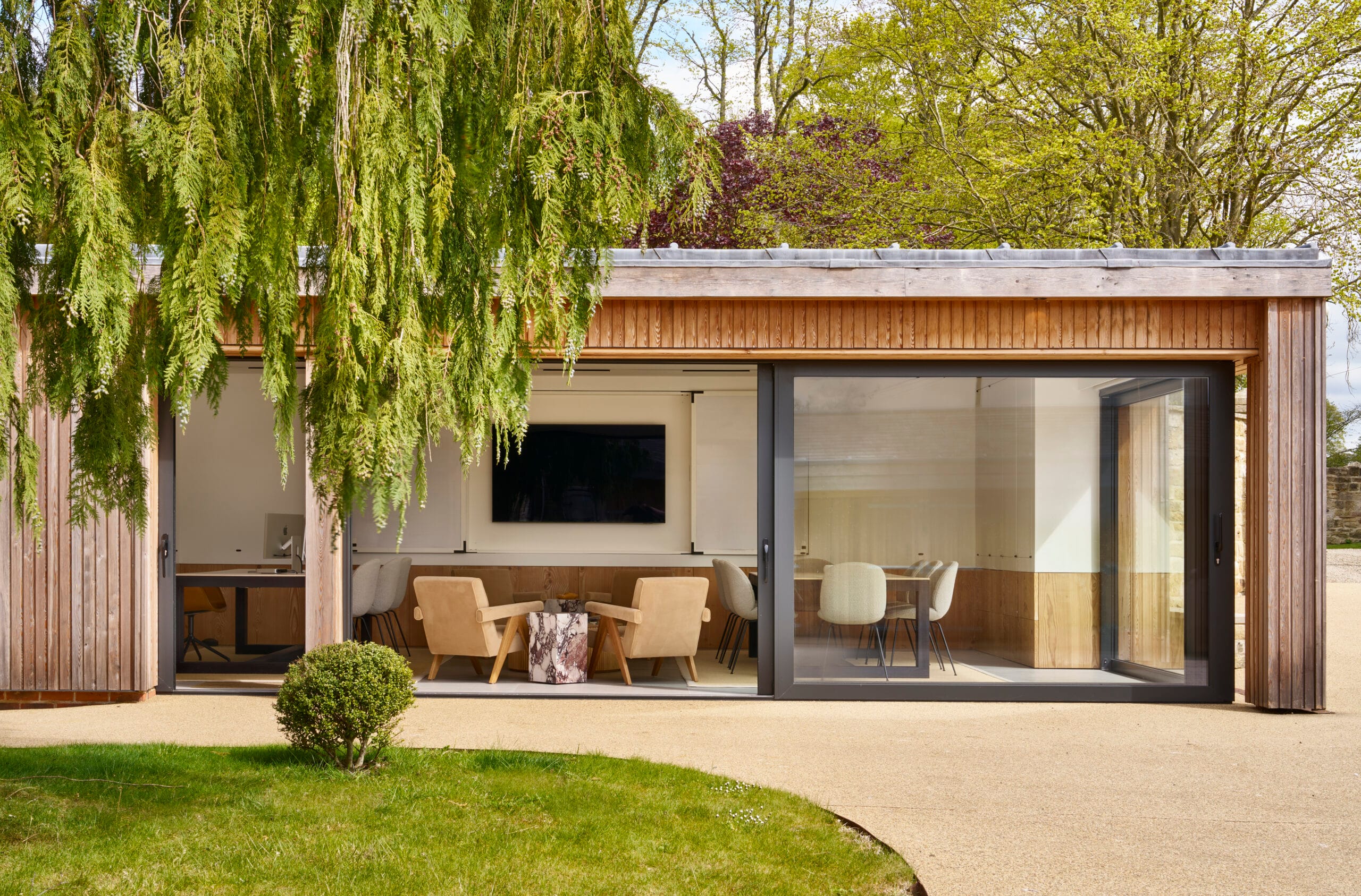 A modern garden office with wooden cladding and large sliding glass doors opening onto a patio. Inside, neutral-toned lounge chairs surround a marble side table, with additional seating and a desk visible in the background. Greenery and trees frame the view.