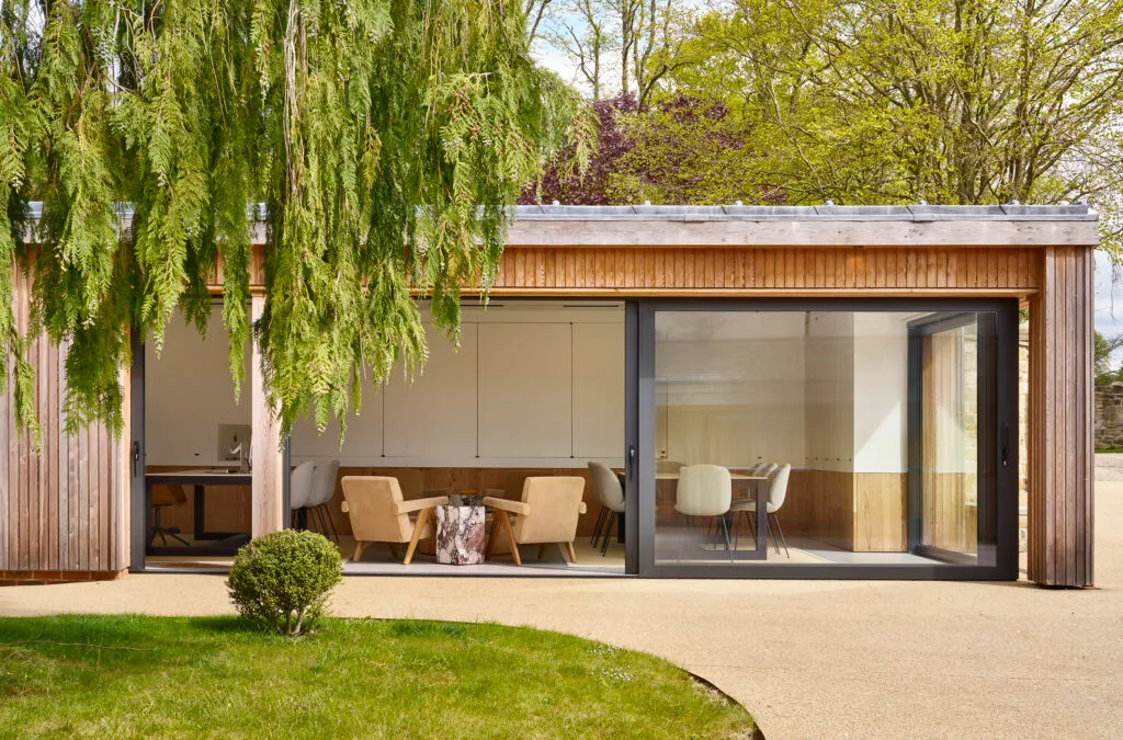 A modern garden office with wooden cladding and large sliding glass doors opening onto a patio. Inside, neutral-toned lounge chairs surround a marble side table, with additional seating and a desk visible in the background. Greenery and trees frame the view.