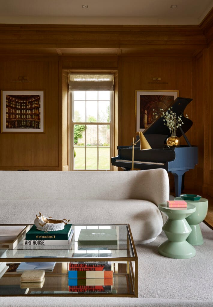A stylish living room featuring wood-paneled walls, a cream sofa, and a glass coffee table with art and architecture books stacked underneath. A blue grand piano stands in the corner near a window with garden views, flanked by framed photographs and a gold decorative vase with flowers.