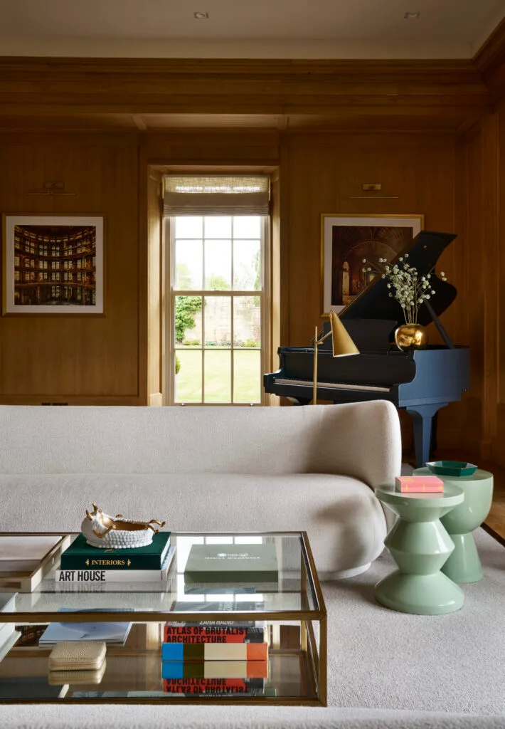 A stylish living room featuring wood-paneled walls, a cream sofa, and a glass coffee table with art and architecture books stacked underneath. A blue grand piano stands in the corner near a window with garden views, flanked by framed photographs and a gold decorative vase with flowers.