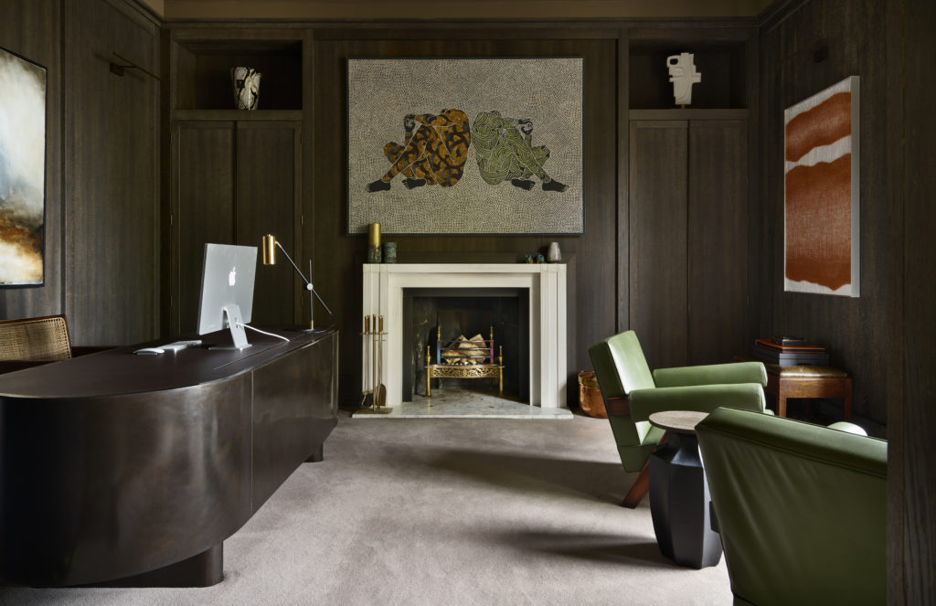 A sophisticated home office featuring dark wood-paneled walls, a modern dark brown curved desk with an Apple iMac, and a sleek brass desk lamp. The room includes a central white fireplace with a marble hearth, above which hangs a large contemporary artwork of two abstract human figures. On either side of the fireplace are built-in cabinets with decorative sculptures. Two green leather armchairs and a black geometric side table create a cozy seating area. Modern art pieces adorn the walls, contributing to the room’s refined and artistic ambiance.