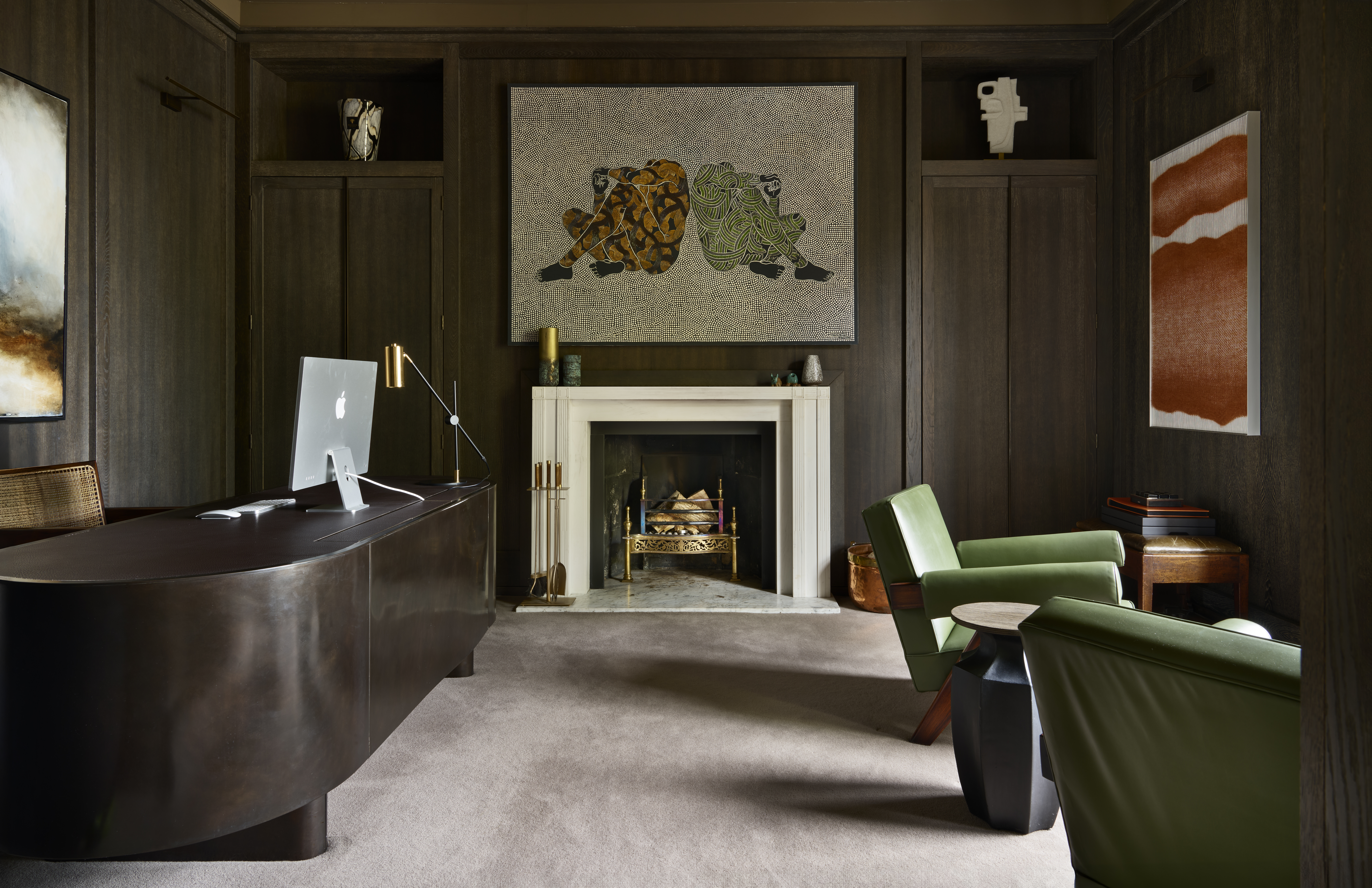 A sophisticated home office featuring dark wood-paneled walls, a modern dark brown curved desk with an Apple iMac, and a sleek brass desk lamp. The room includes a central white fireplace with a marble hearth, above which hangs a large contemporary artwork of two abstract human figures. On either side of the fireplace are built-in cabinets with decorative sculptures. Two green leather armchairs and a black geometric side table create a cozy seating area. Modern art pieces adorn the walls, contributing to the room’s refined and artistic ambiance.