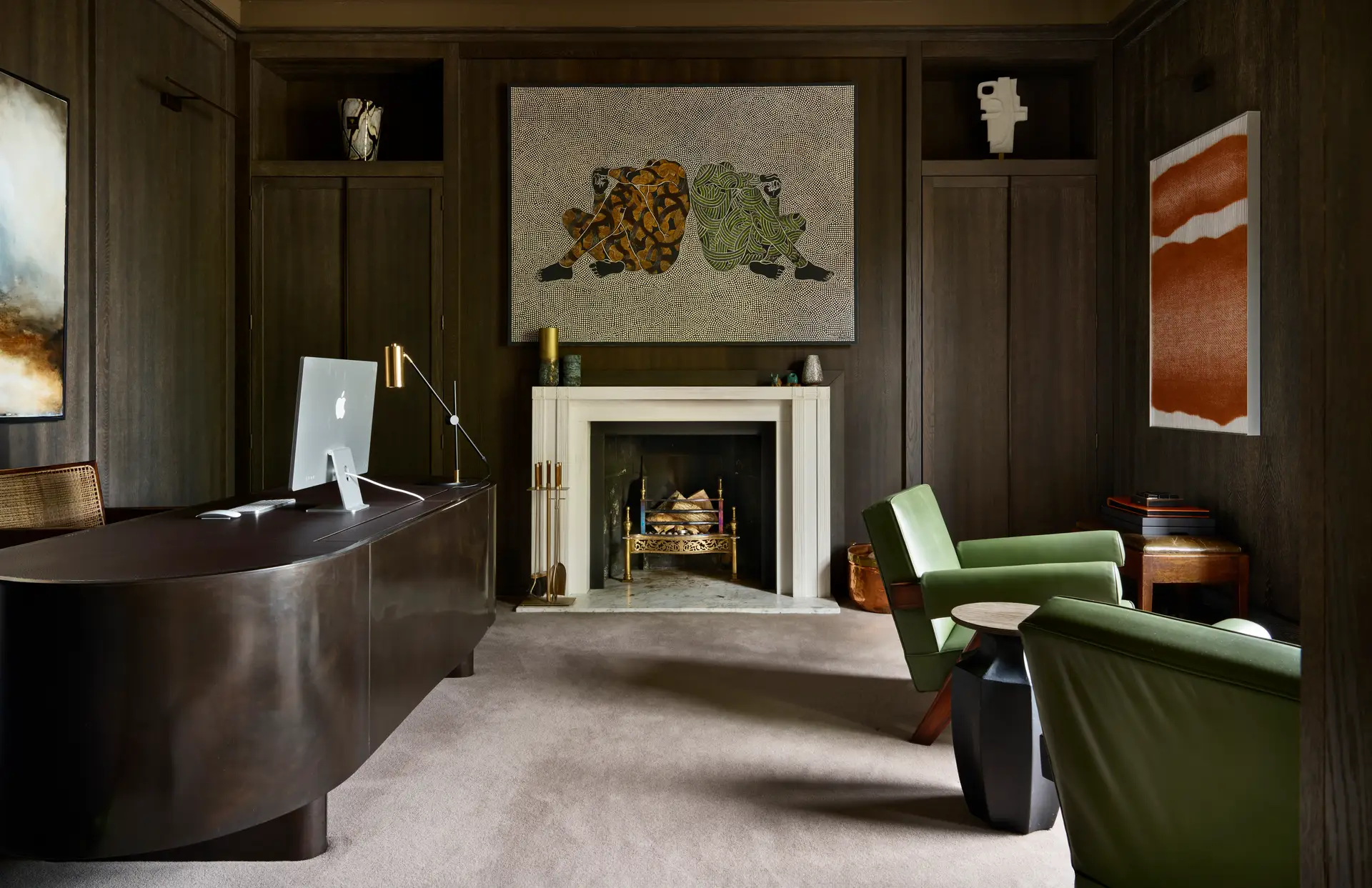A sophisticated home office featuring dark wood-paneled walls, a modern dark brown curved desk with an Apple iMac, and a sleek brass desk lamp. The room includes a central white fireplace with a marble hearth, above which hangs a large contemporary artwork of two abstract human figures. On either side of the fireplace are built-in cabinets with decorative sculptures. Two green leather armchairs and a black geometric side table create a cozy seating area. Modern art pieces adorn the walls, contributing to the room’s refined and artistic ambiance.