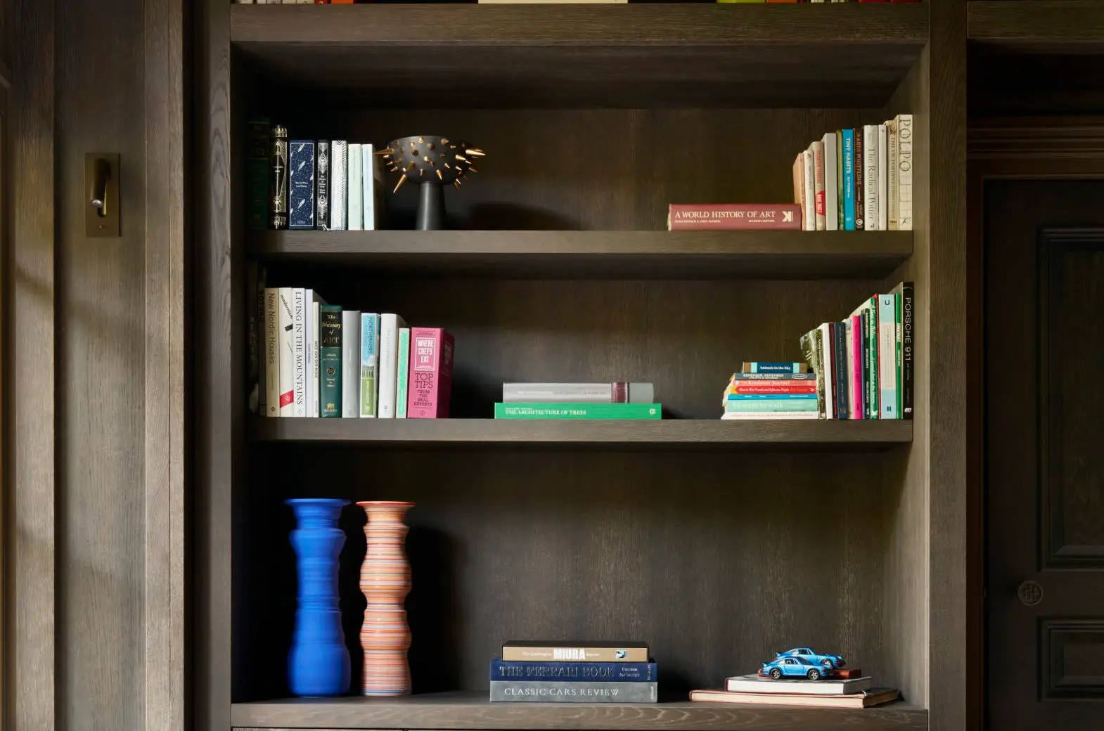 A cozy reading nook with a dark wooden built-in bookshelf filled with colorful books and decorative objects, including sculptural vases and a small model car. A green mid-century modern armchair sits in front, and natural light enters through a nearby window.
