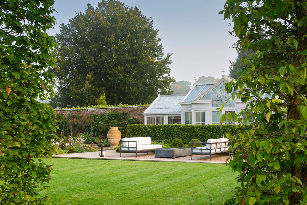 Walled garden with greenhouse and neat hedge, seating area, lawn, and shrubbery in the foreground.